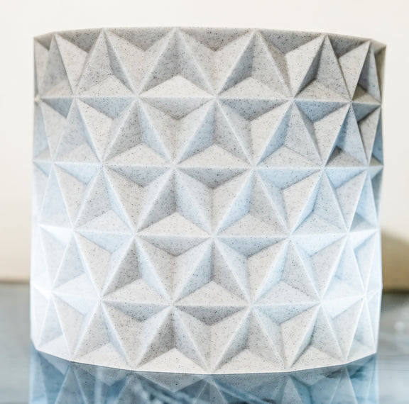 A cylindrical planter with a geometric, faceted 3D star pattern, made from light gray speckled concrete or stone, shown on a reflective surface against a neutral background.